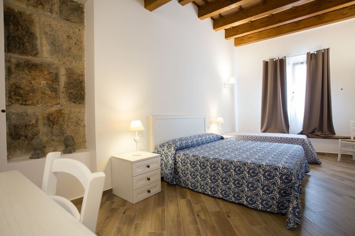 Quarto com parede de pedra, vigas de madeira, cama com roupa de cama estampada, luminárias de cabeceira e janela com cortinas.