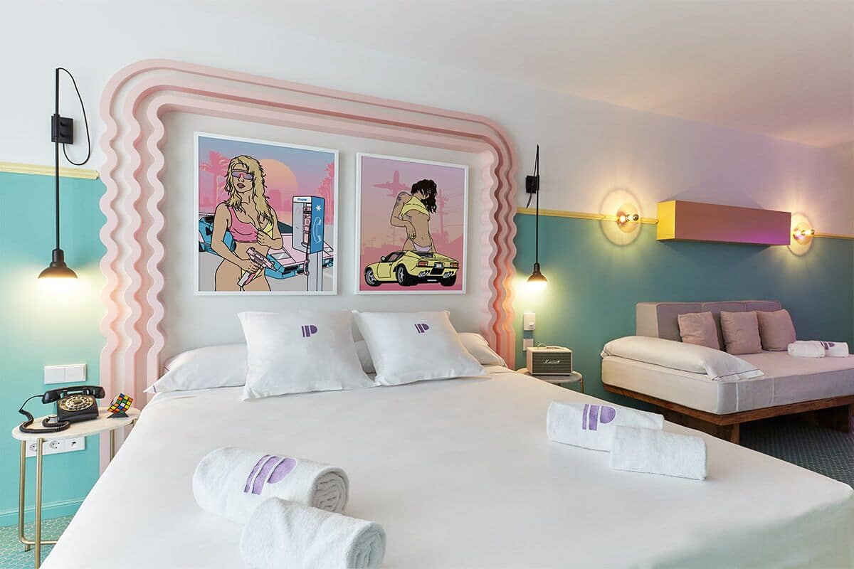 Ett hotellrum med en säng, två väggmonterade lampor, ett sängbord med en telefon och två inramade konstverk.
