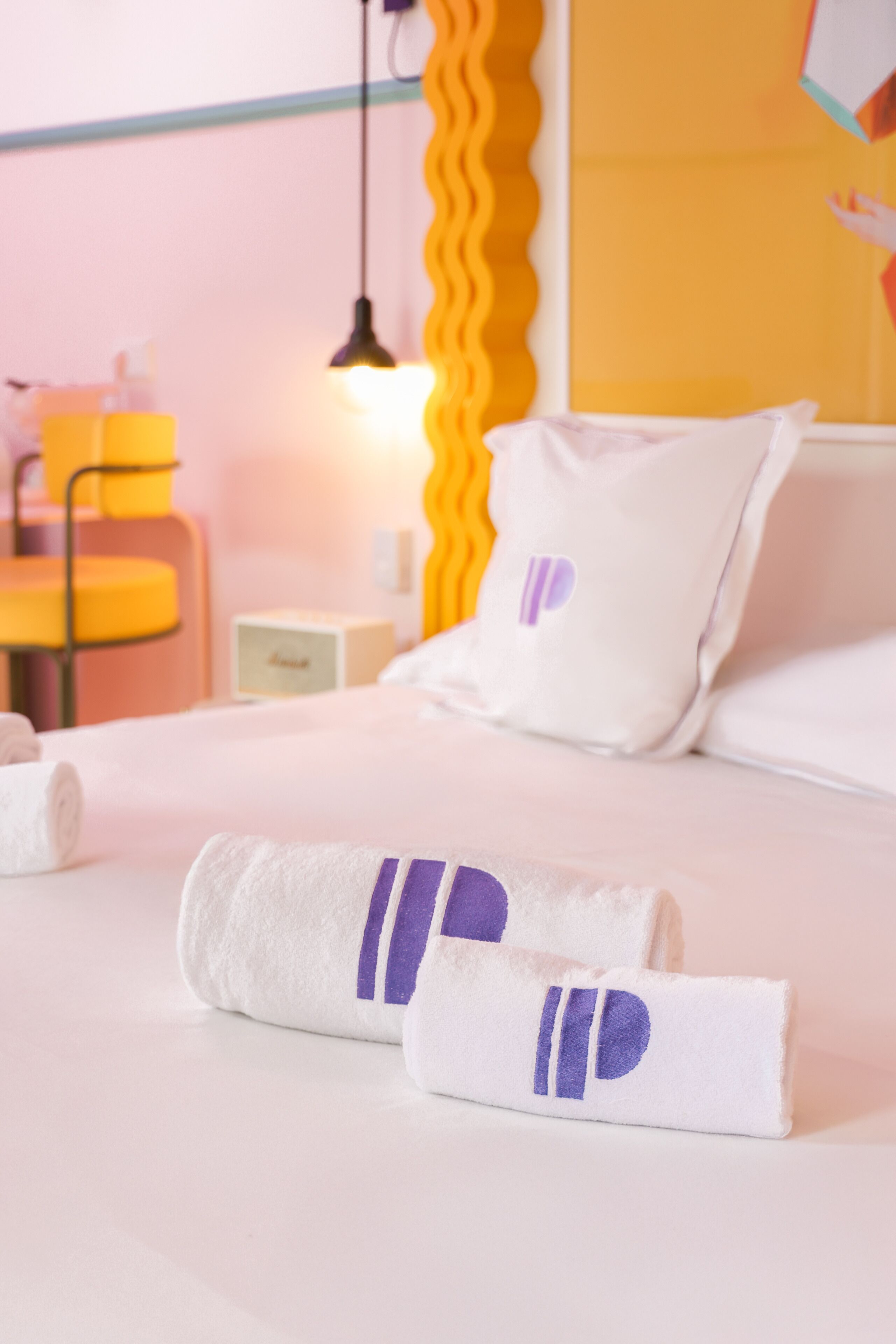 Un lit bien fait, avec du linge de lit blanc et des touches de couleur violette, une table de chevet avec une lampe et un miroir jaune aux motifs ondulés.