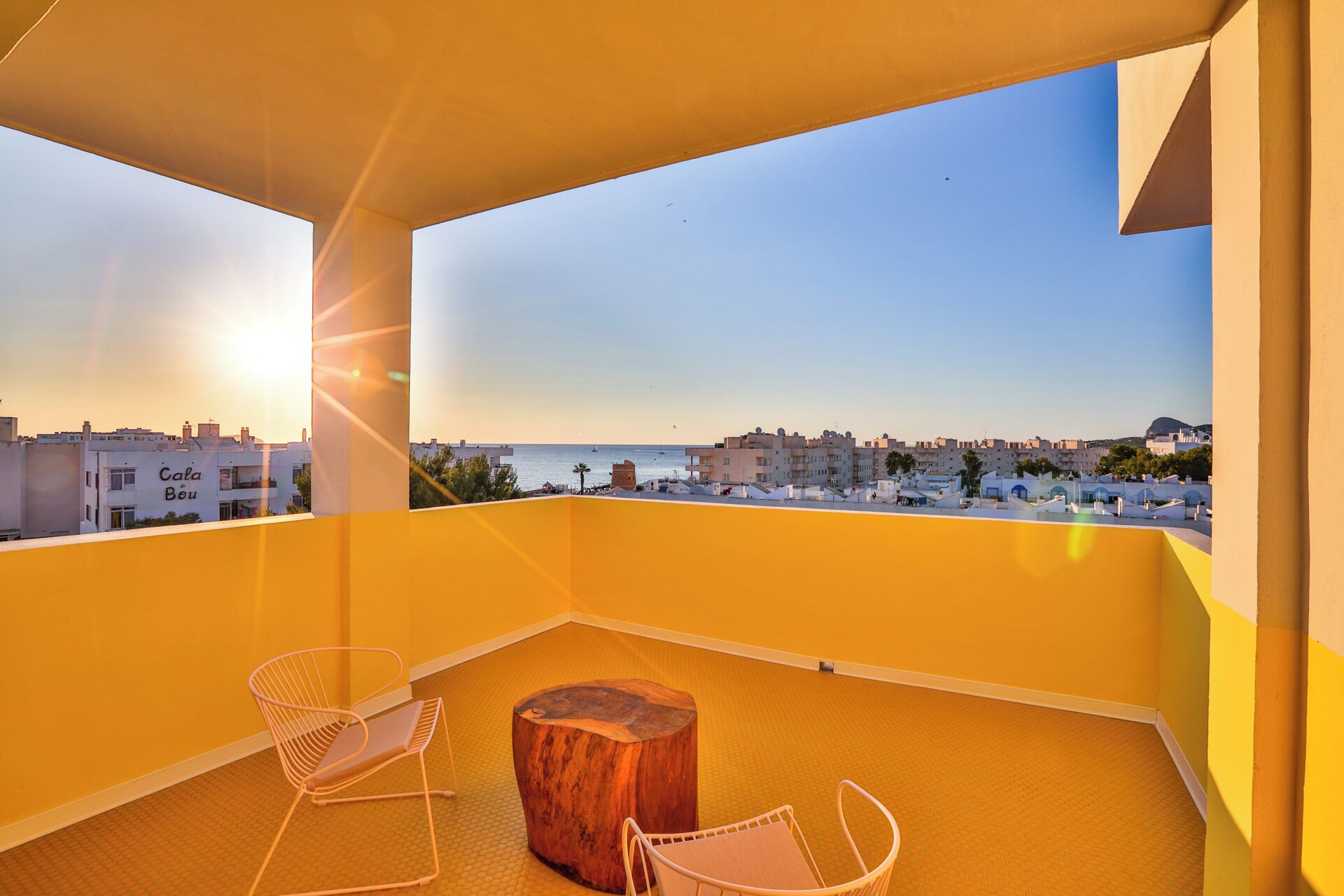 A balcony with two chairs and a wooden stool, overlooking a coastal cityscape during sunset.