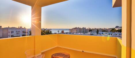 A balcony with two chairs and a wooden stool, overlooking a coastal cityscape during sunset.