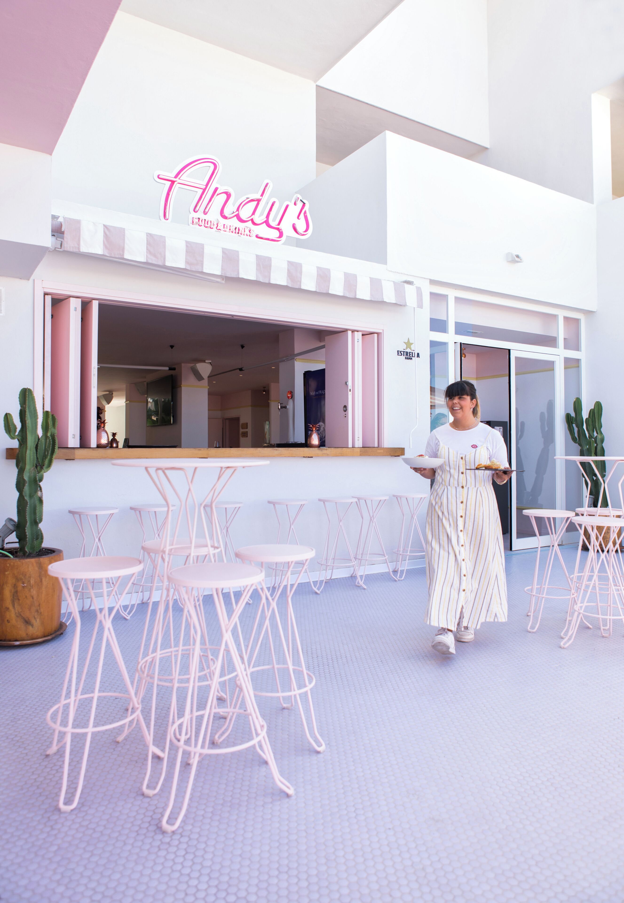 Zona salotto all'aperto moderna con sedie e tavoli rosa, una pianta di cactus e un neon con scritto 'Andy's'.