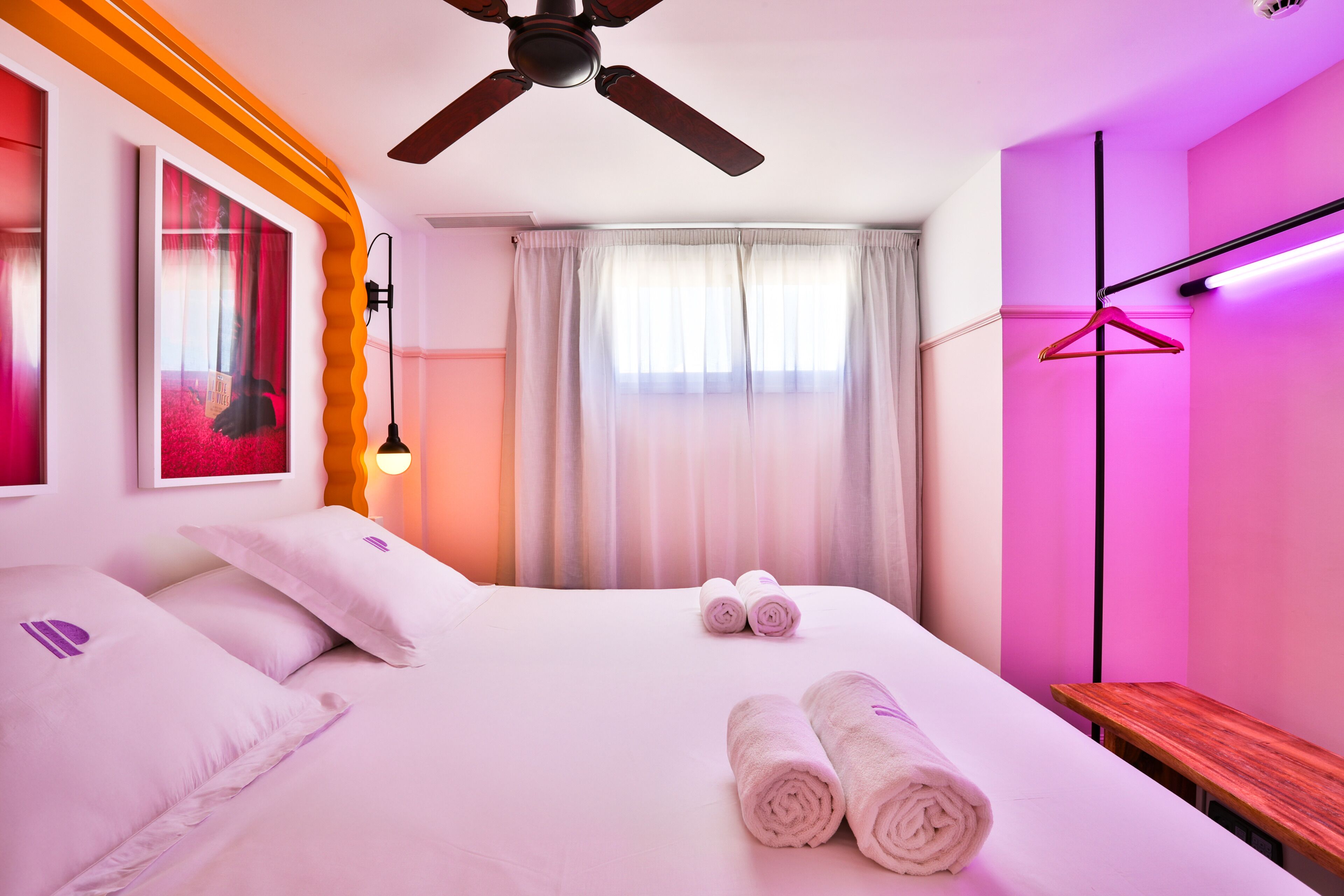 Habitación de hotel con cama, almohadas, toallas enrolladas, ventilador de techo, ventana con cortinas, mesita de noche y lámpara de pared.