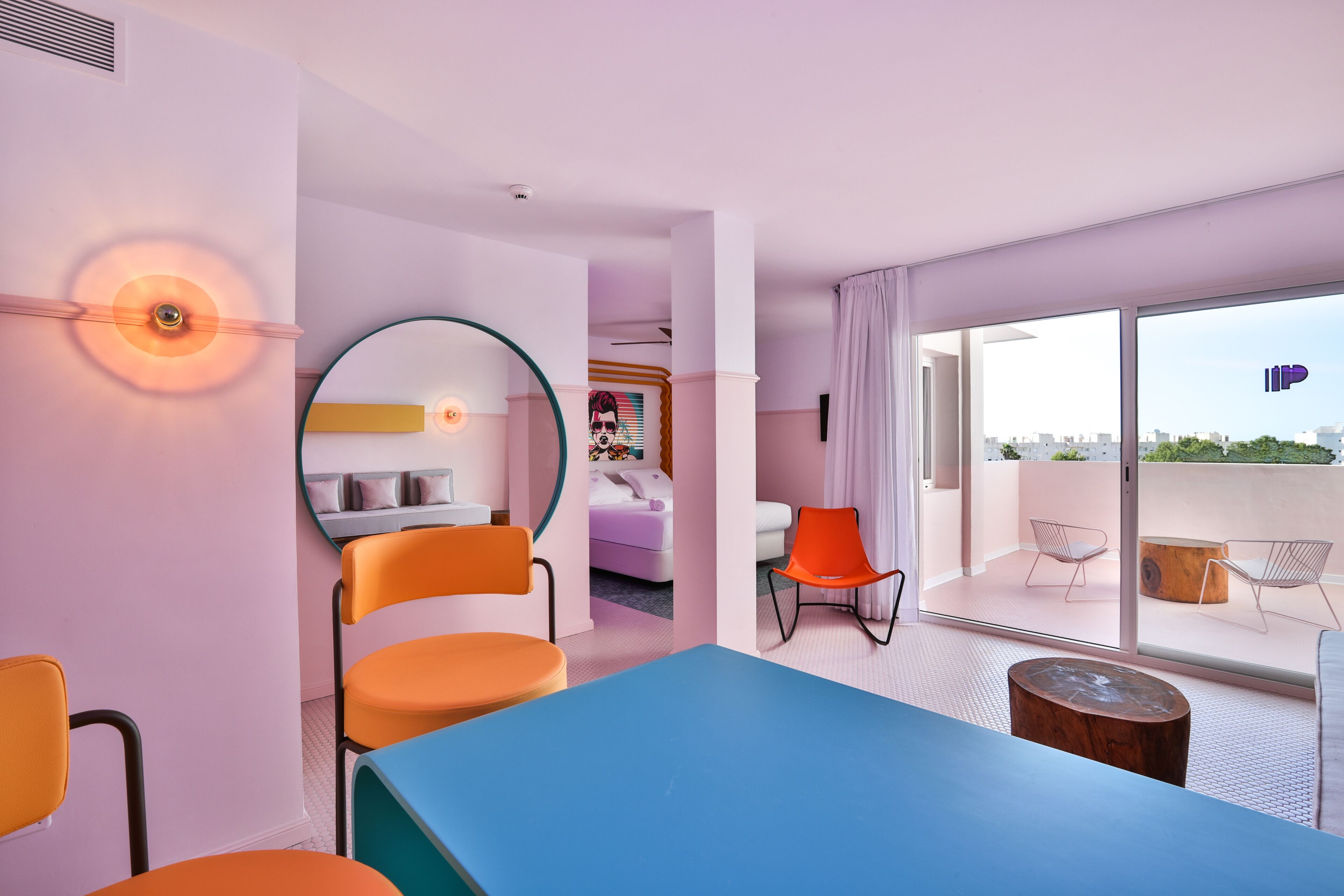 A modern hotel room with a large window, a round mirror, a blue table, and orange chairs.