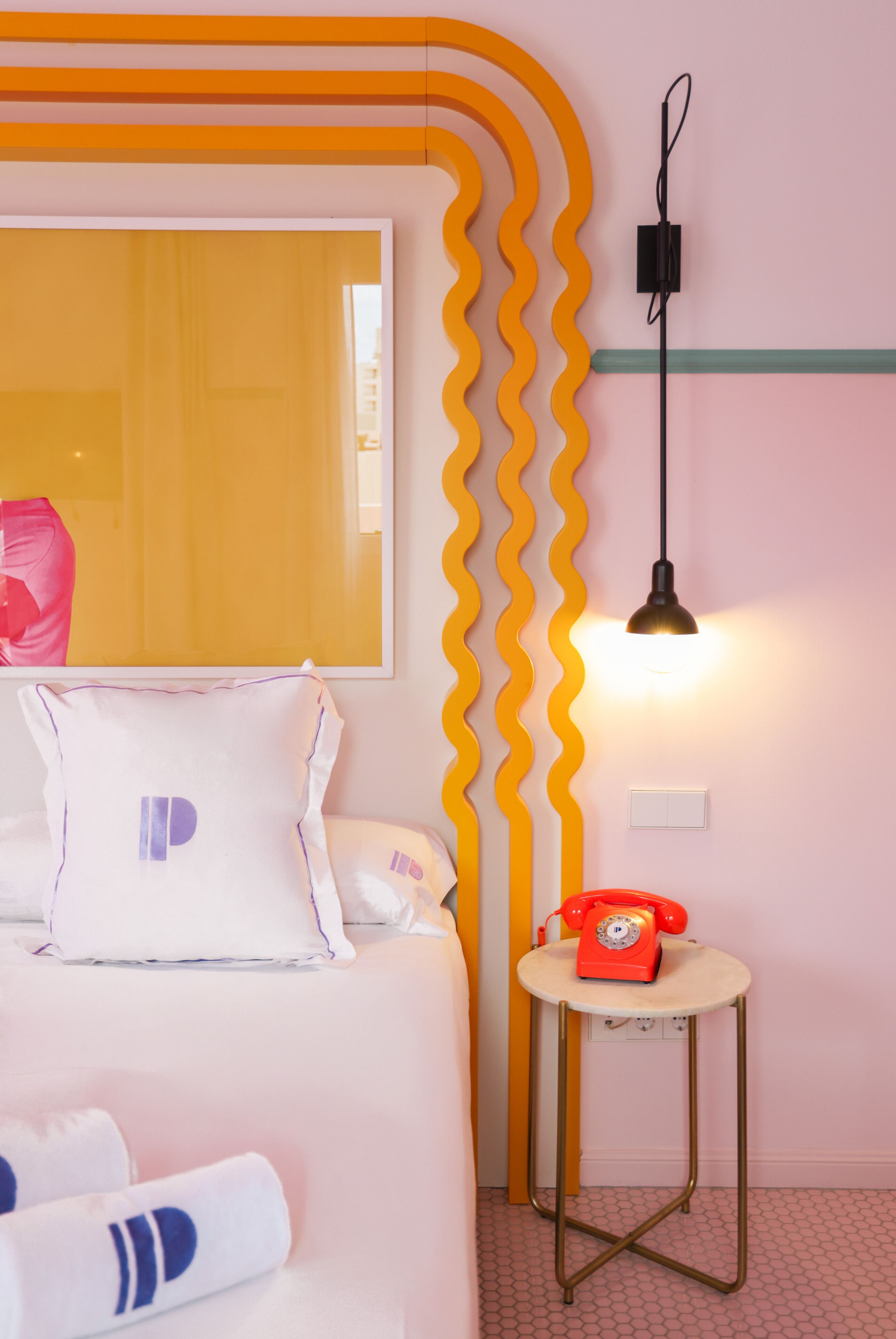 Een hotelkamer met een bed, kussens, een rode telefoon, een wandlamp en een spiegel.