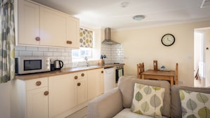 Cocina compacta con microondas, hervidor y lavabo. Hay una mesa de comedor con sillas y un reloj en la pared.
