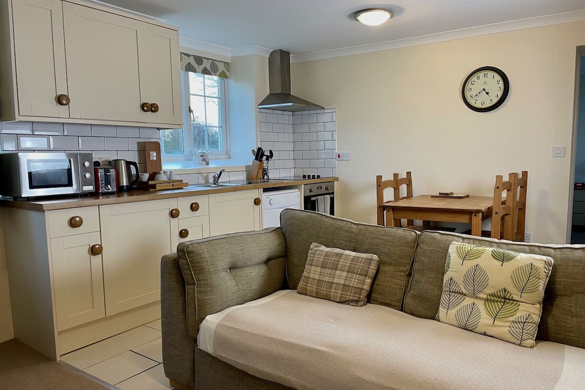 A compact kitchen with a microwave, toaster, kettle, and stove. There's a dining area with a wooden table and chairs, and a sofa with patterned cushions.
