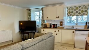 Cocina compacta con armarios blancos, microondas, tostadora y lavavajillas. En la zona de estar, hay un sofá gris, un televisor y una ventana con cortinas.