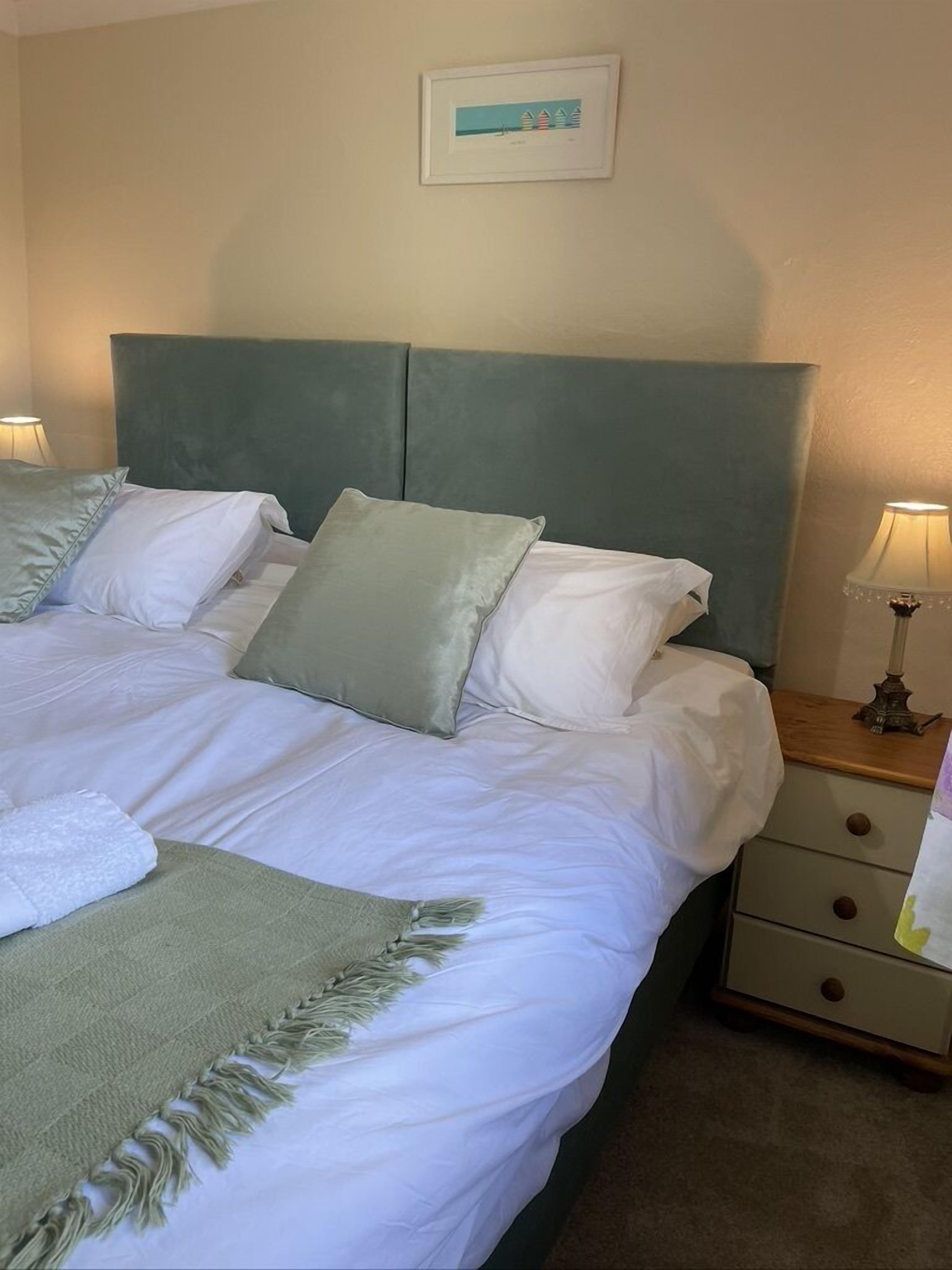 A neatly made bed with white linens and a green throw blanket, a bedside table with a lamp, and a wall-mounted picture.