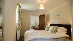 Una cama bien hecha con sábanas blancas y algunos almohadones, una mesita de noche con una lámpara, y vista a un baño con un perchero para toallas.