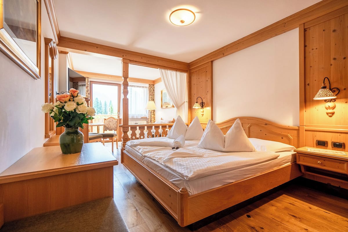 Un dormitorio con estructura de madera, con una cama, un escritorio y una silla. Hay una ventana con cortinas, una lámpara y un florero con flores.