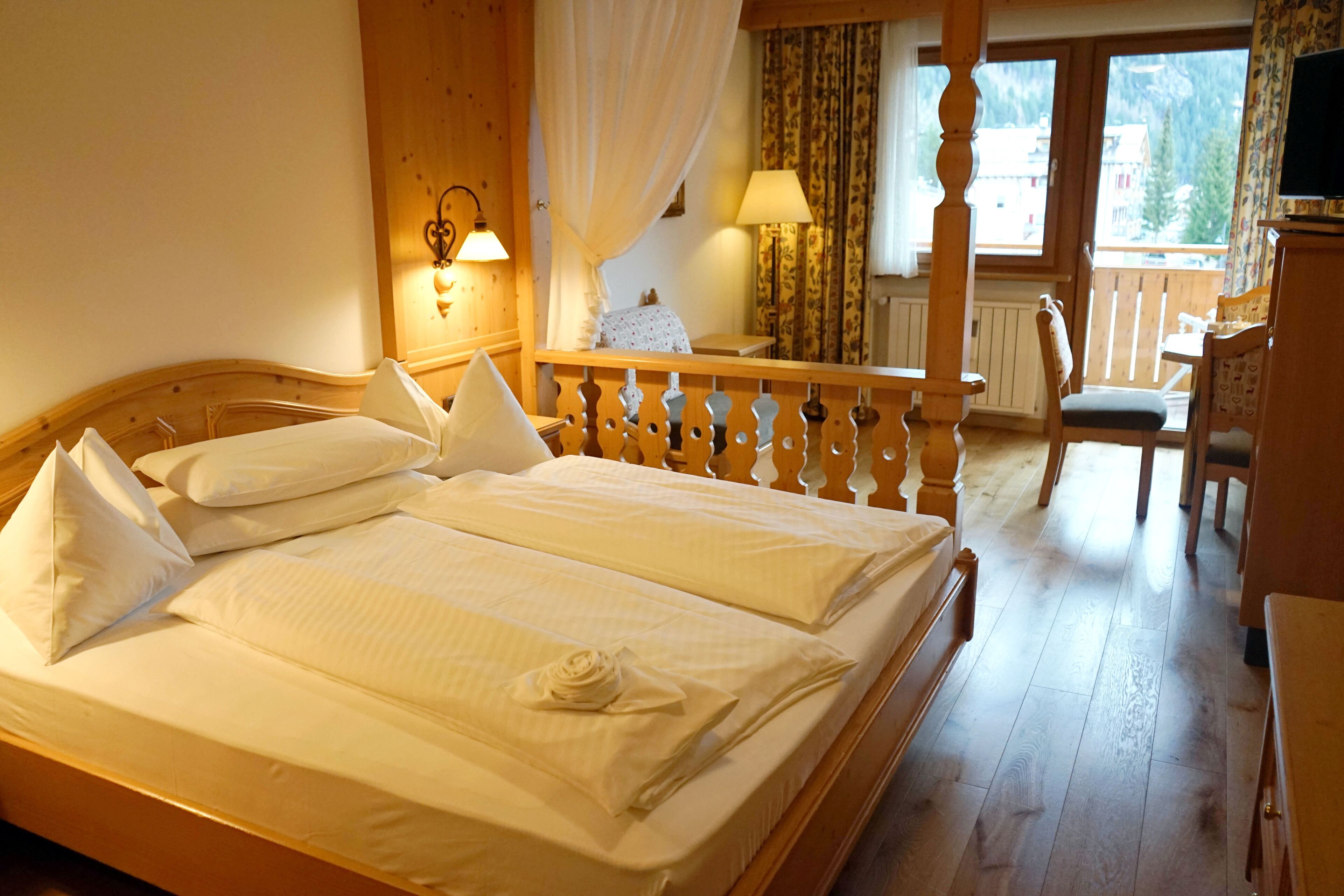 A double bed with white linens and pillows, a wooden headboard, a nightstand with a lamp, a wooden chair, and a view of a mountain through a window.