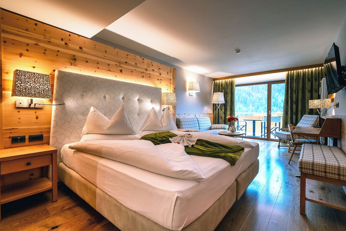 Habitación de hotel con una cama grande, cabecero de madera, escritorio con silla y balcón con vistas al exterior.