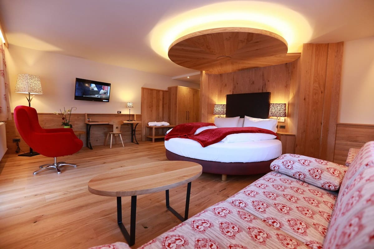 Habitación de hotel moderna con techo de madera, cama redonda, sillón rojo, sofá, escritorio de madera y televisor en la pared.