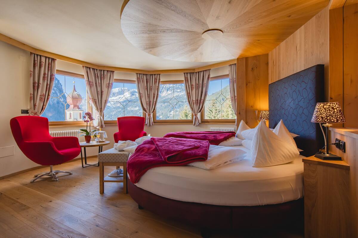 Un dormitorio con una cama grande, dos sillas rojas, una mesita y vistas a las montañas a través de amplios ventanales.