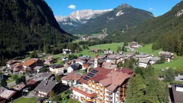 A town with a prominent building featuring balconies and solar panels, surrounded by lush greenery and mountains in the background.