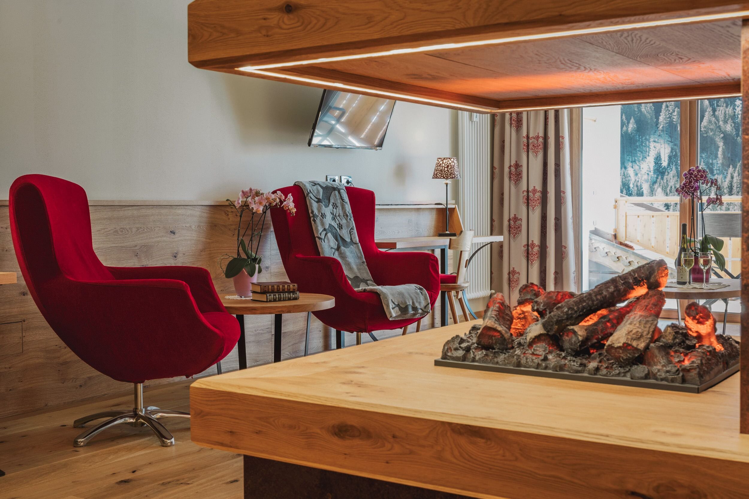 Ein gemütliches Zimmer mit Holzbalkendecke, roten Stühlen, einem kleinen Tisch mit Pflanze und einem Kamin mit Holzscheiten.