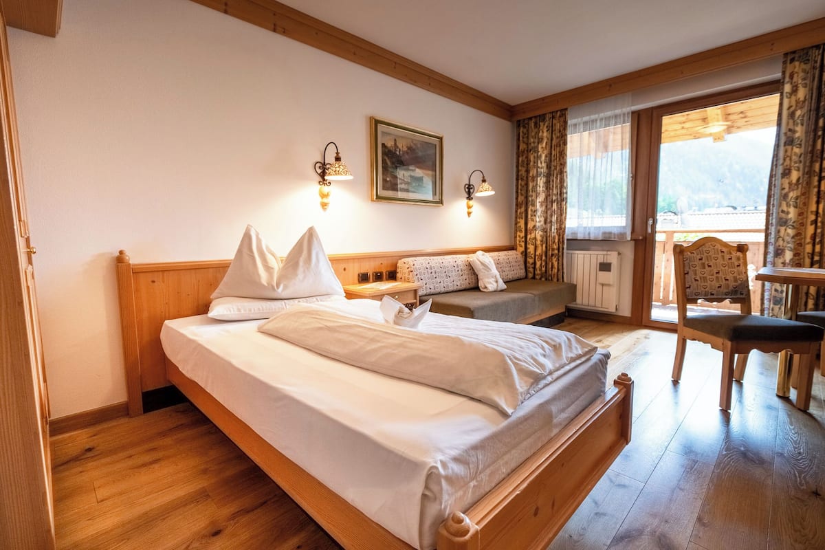 Una habitación de hotel con una cama de madera, dos mesitas de noche, una silla, una ventana con cortinas y un cuadro en la pared.