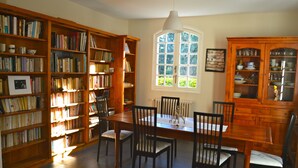 Una biblioteca con estanterías de madera, una mesa de comedor con sillas y un ventanal con vistas a la vegetación.
