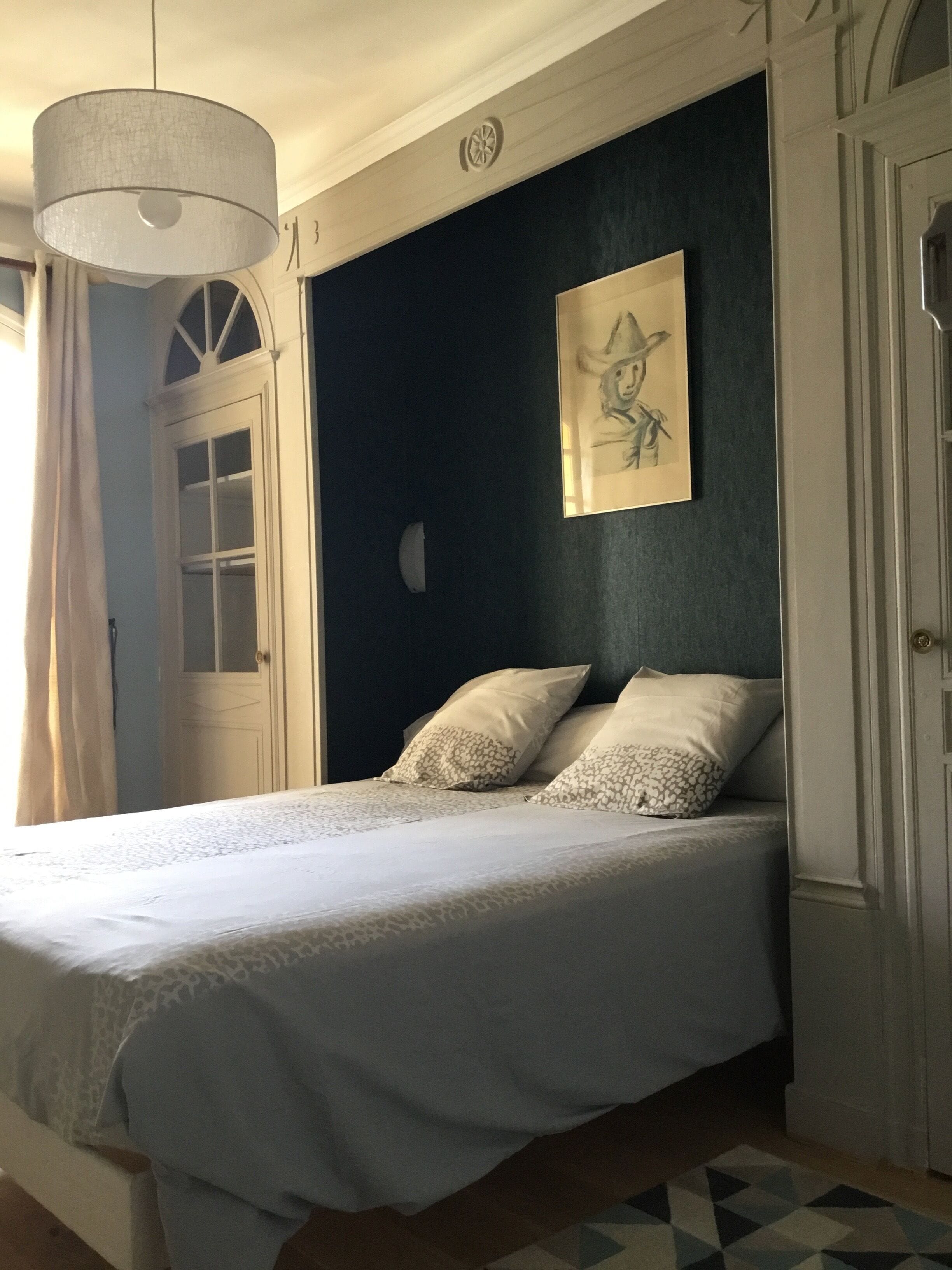 A bedroom with a bed, a large window, a door, and a wall-mounted artwork.