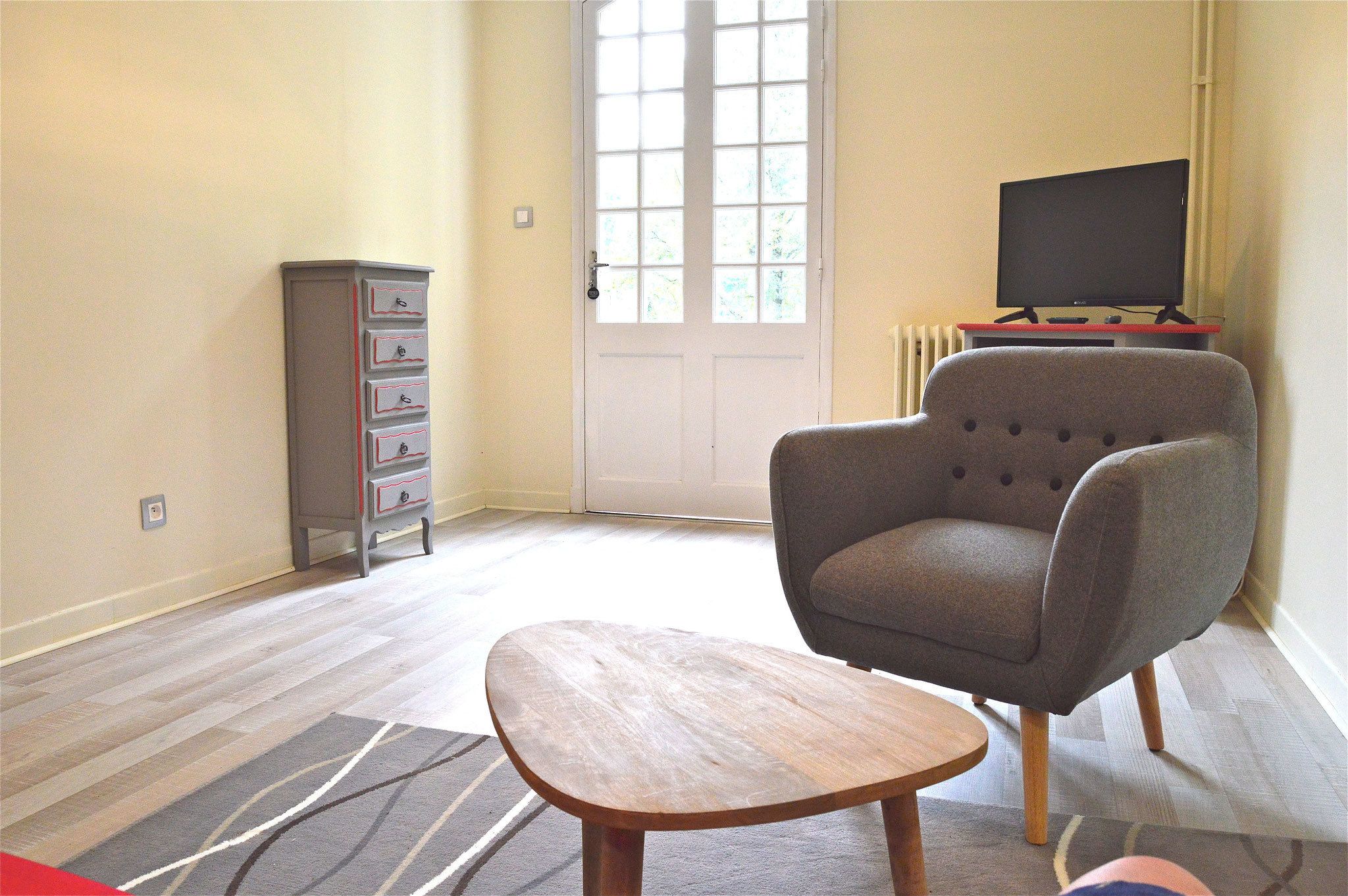 Un salon avec un fauteuil gris, une petite table en bois et un téléviseur posé sur un meuble.