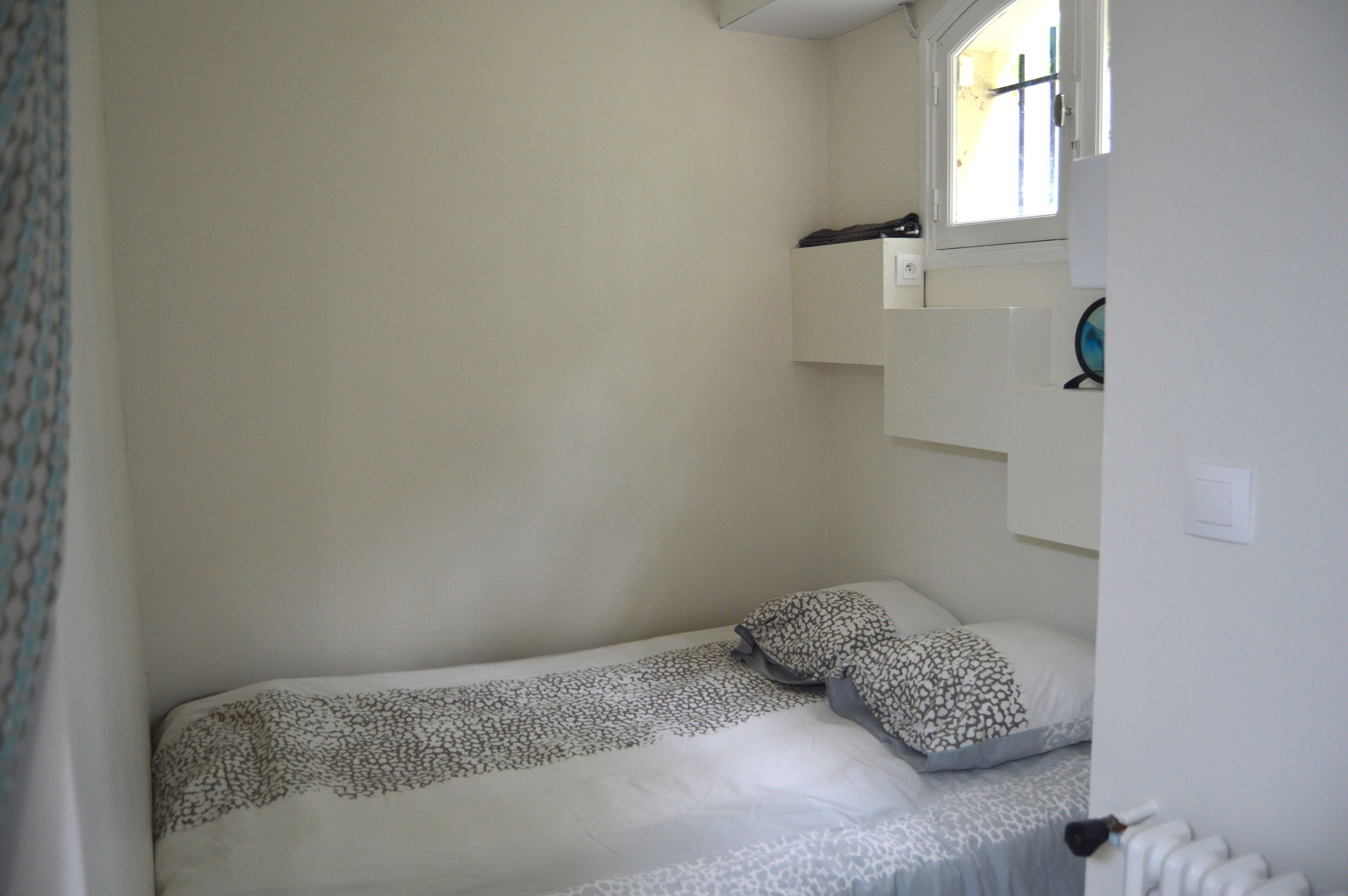 A small, single-bed room with a simple layout, a window, and a small shelf with a globe.