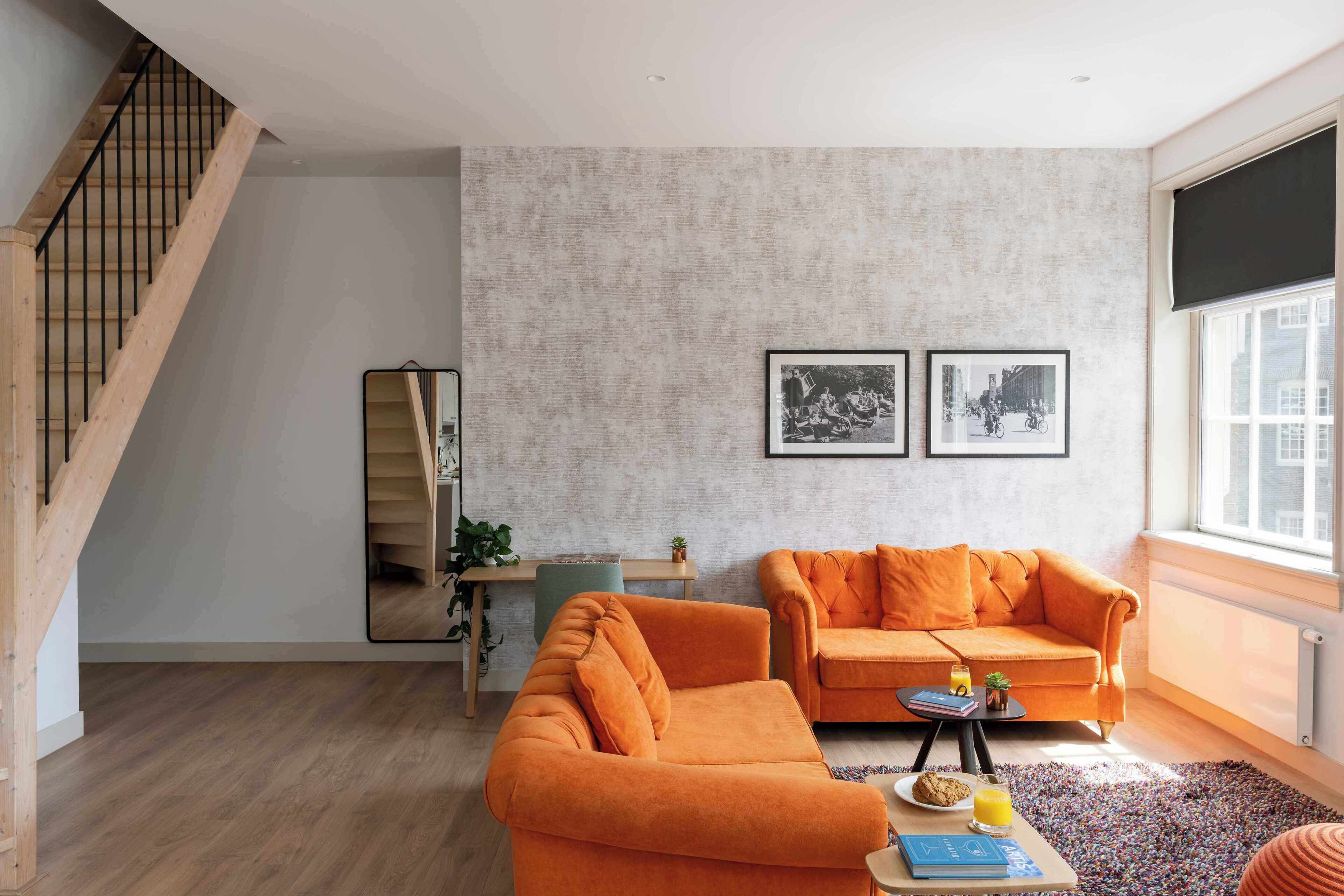 Een moderne woonkamer met een trap, twee oranje banken, een salontafel met boeken en een drankje, en ingelijste foto’s aan de muur.