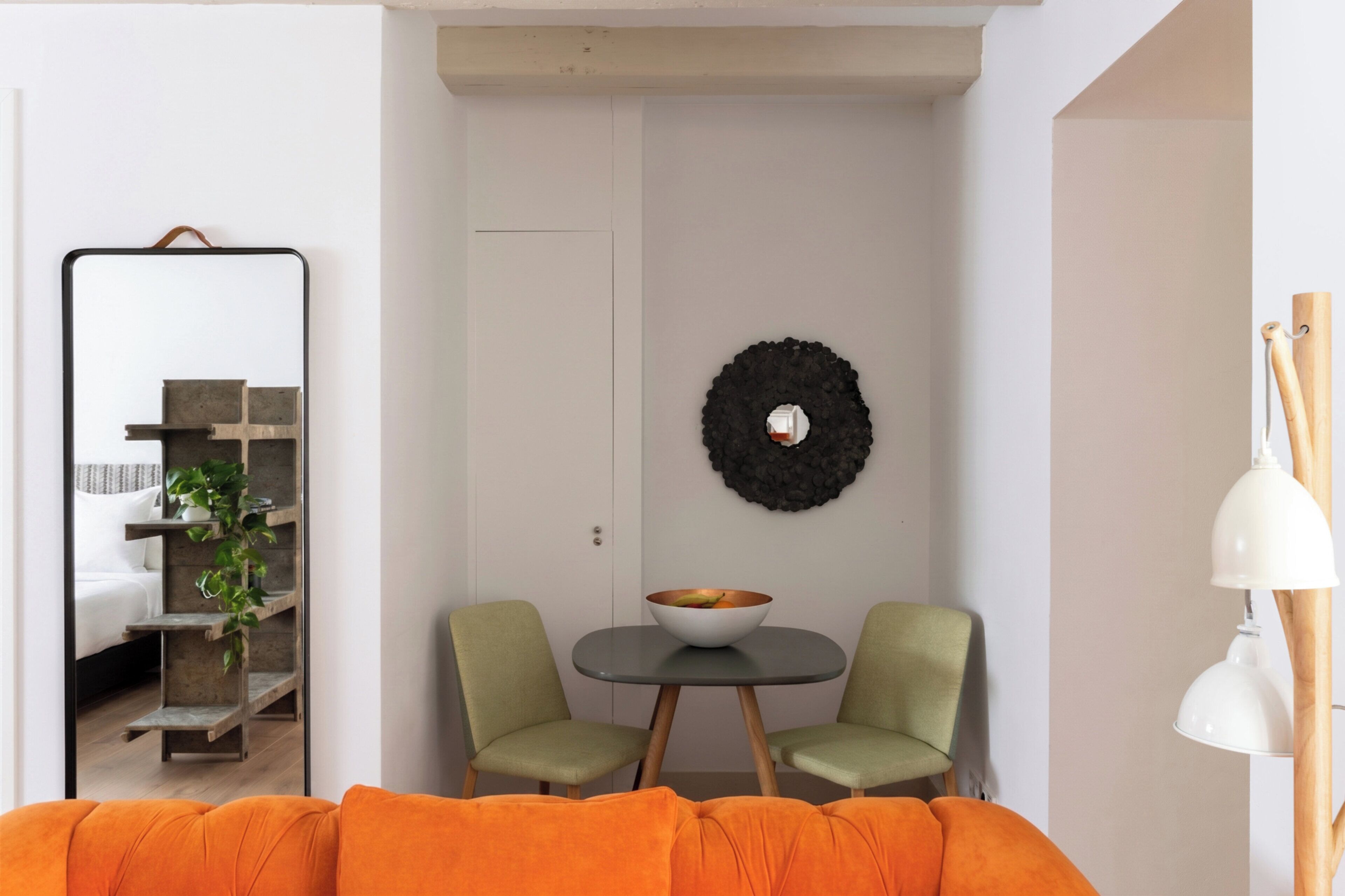A room with a bed, a mirror, a round table with chairs, a wall-mounted light fixture, and a plant.