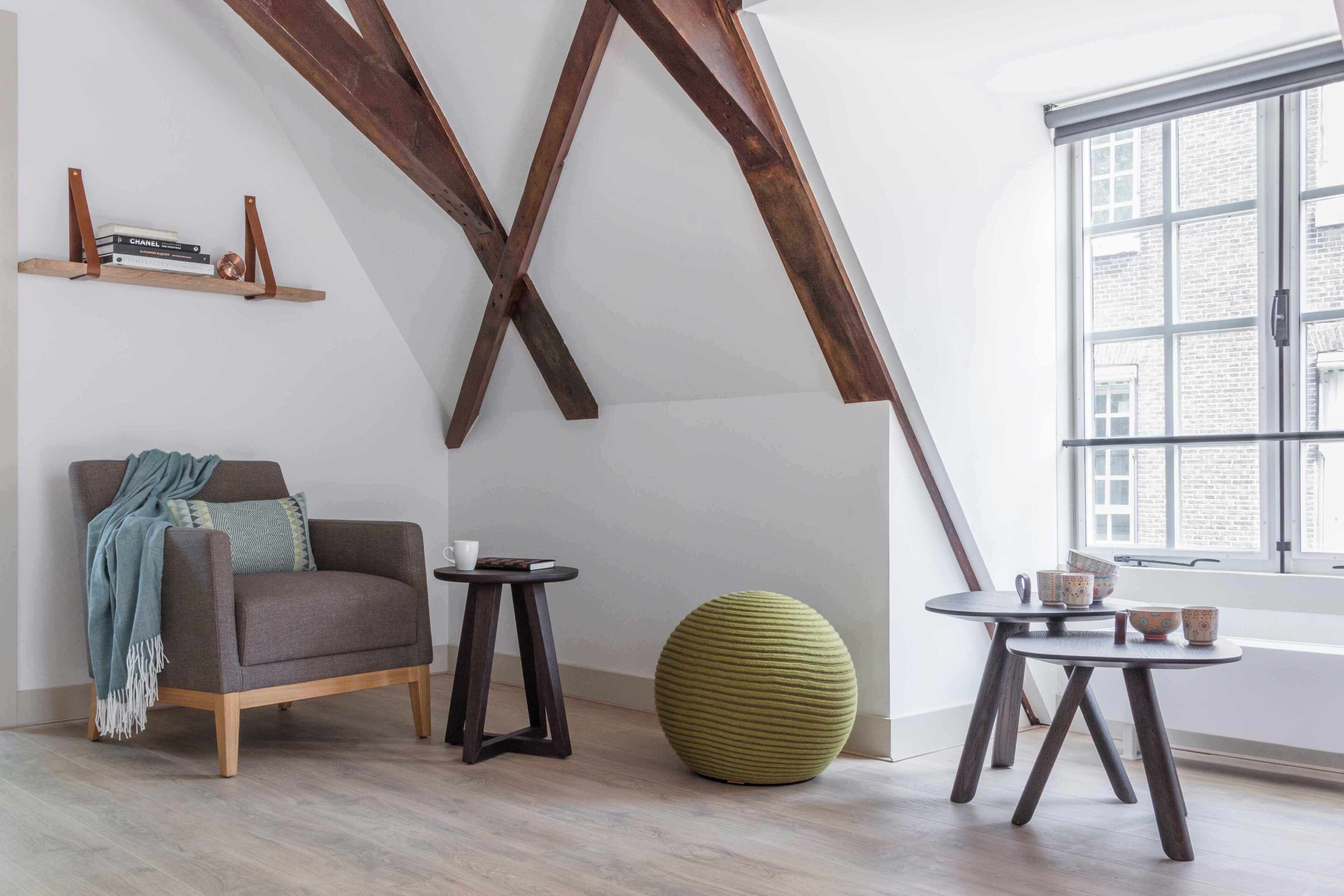A room with a high ceiling, exposed beams, a sofa, a small round table, a round stool, and a window with a view of buildings.
