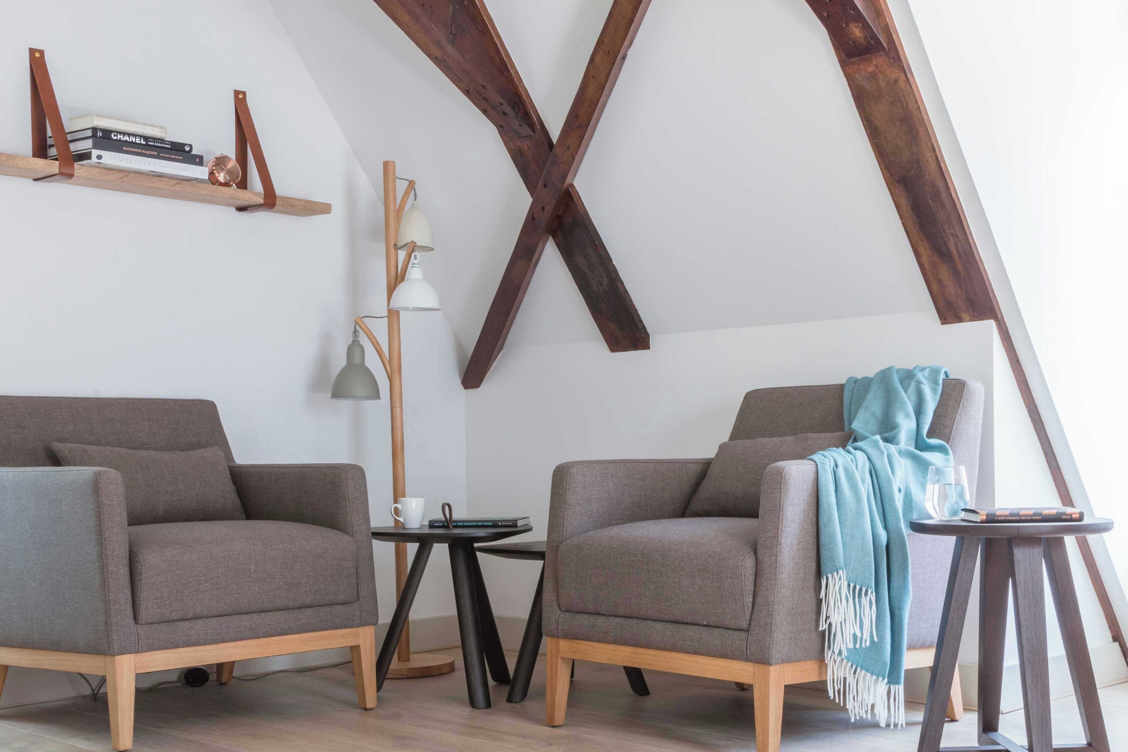 A loft-style room with exposed wooden beams, two grey armchairs, a small round table, and a floor lamp.