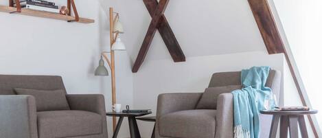 A loft-style room with exposed wooden beams, two grey armchairs, a small round table, and a floor lamp.