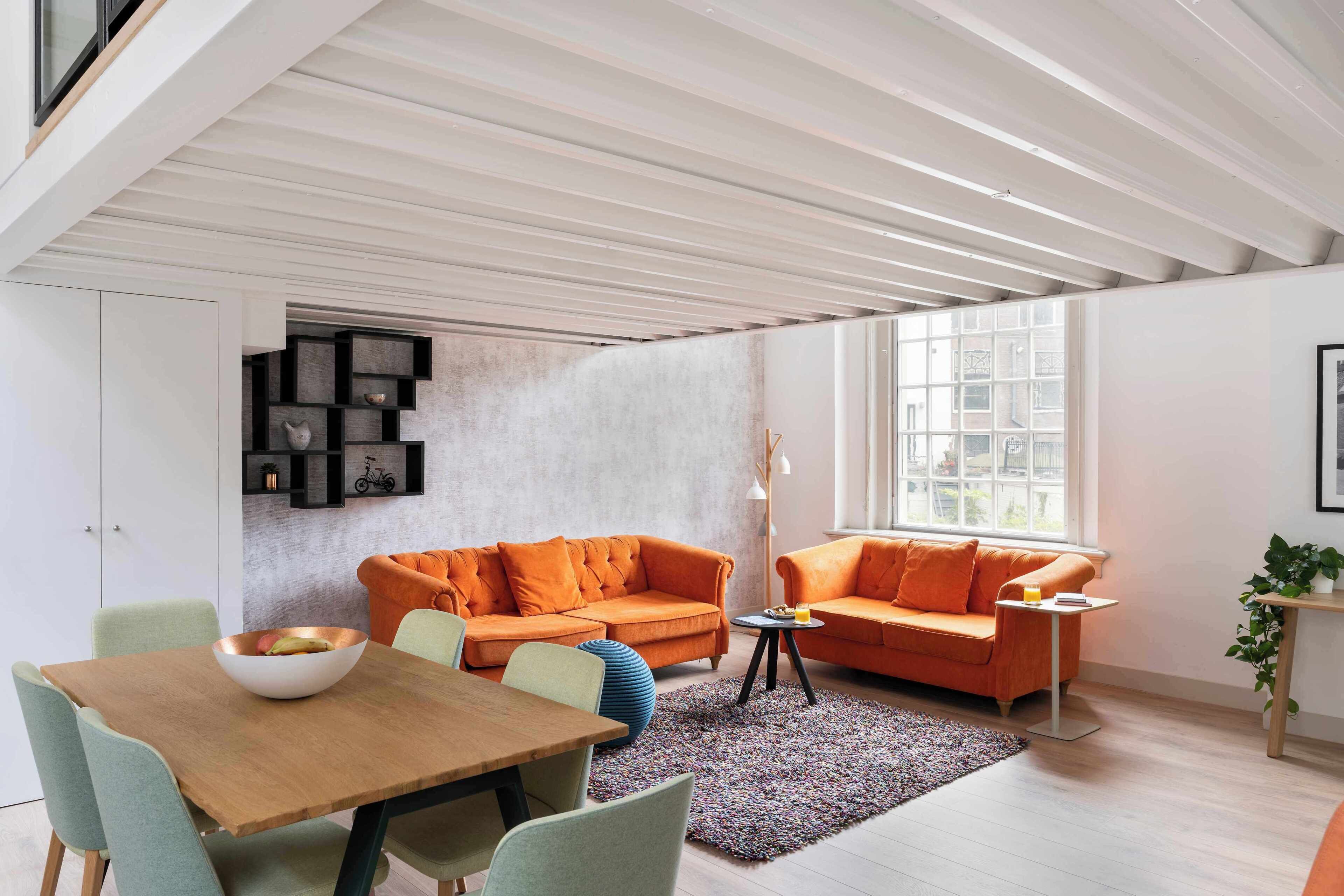 A modern living room with two orange sofas, a wooden dining table, and a large window.