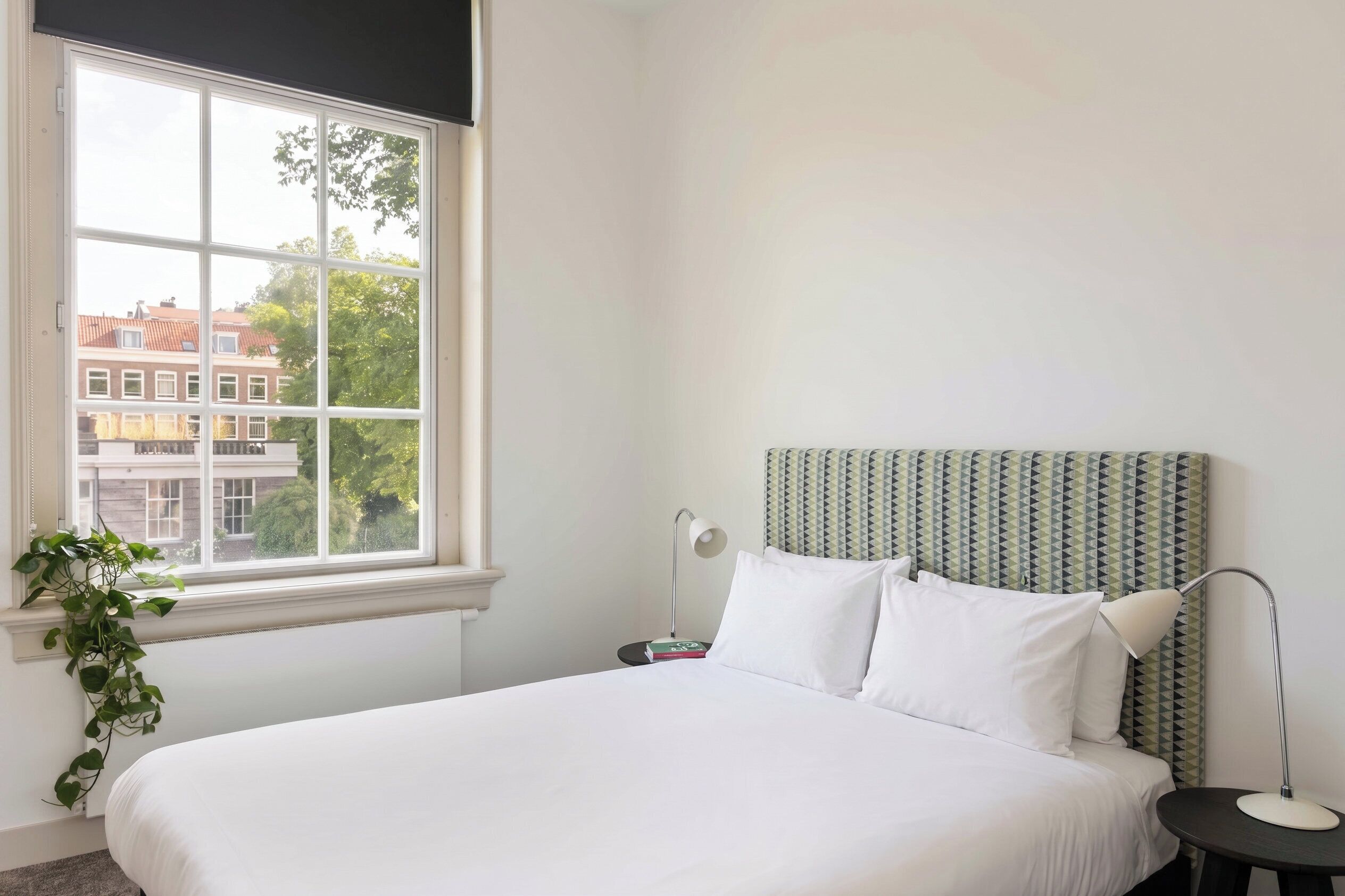 A bedroom with a large window, a bed, a bedside table with a lamp, and a patterned headboard.
