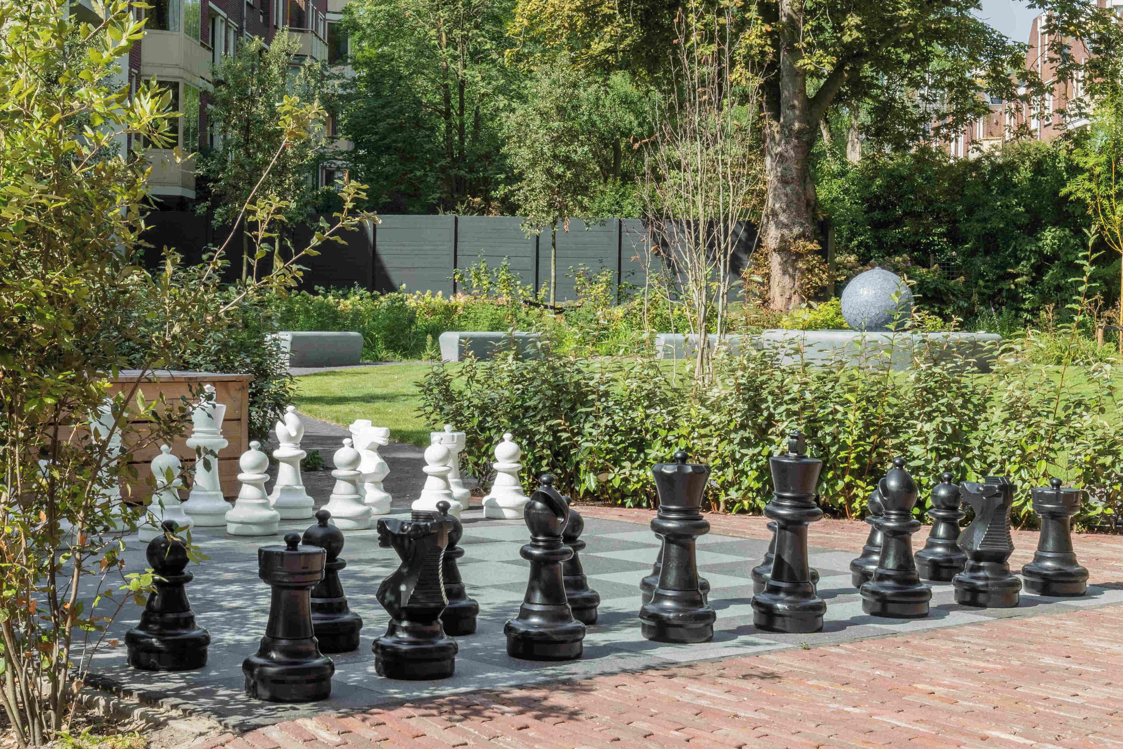 A chessboard with chess pieces set up outdoors, surrounded by greenery and a building in the background.