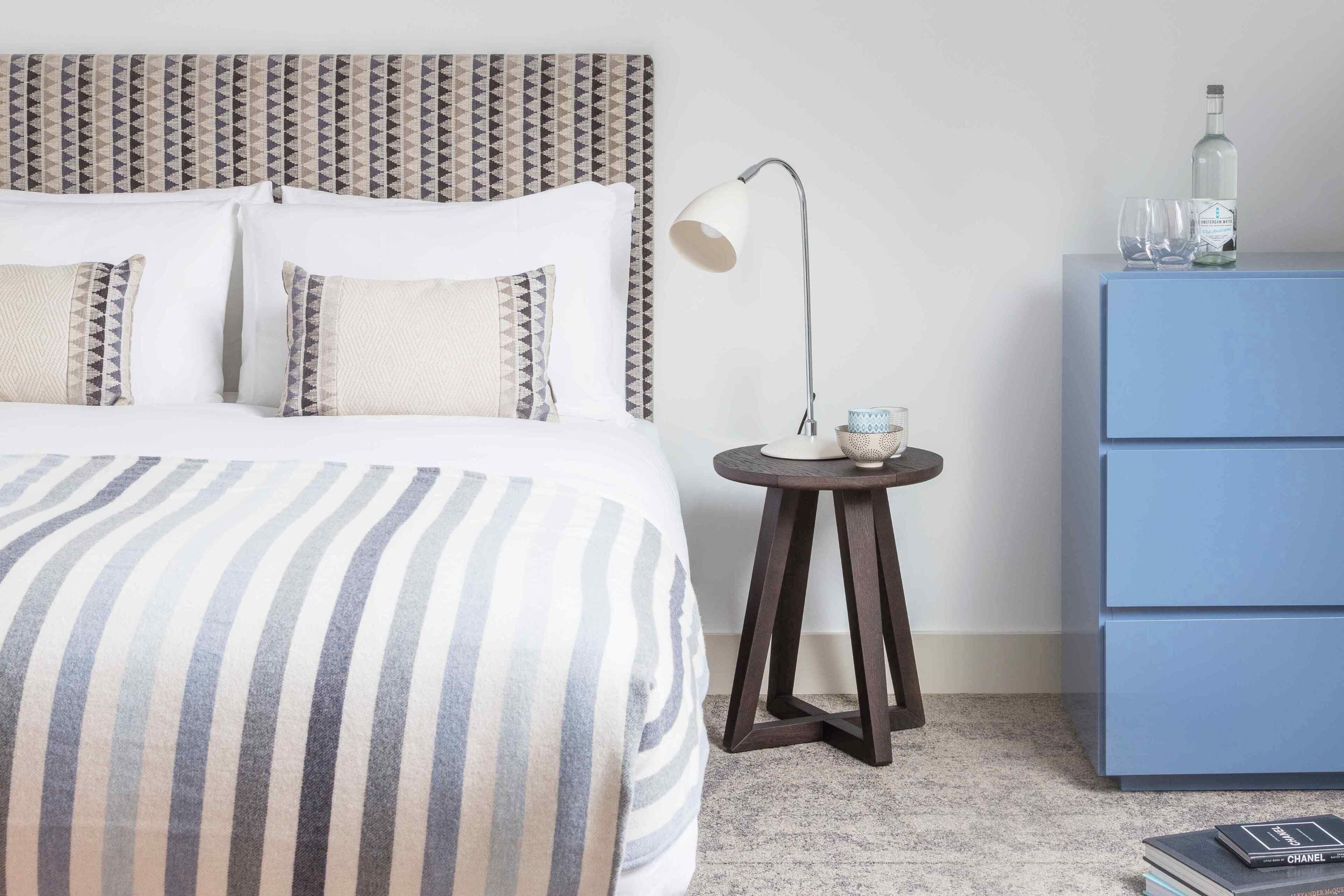 A neatly arranged hotel room with a bed, a bedside lamp, a small round table with a lamp, and a blue dresser with a bottle and glasses on top.