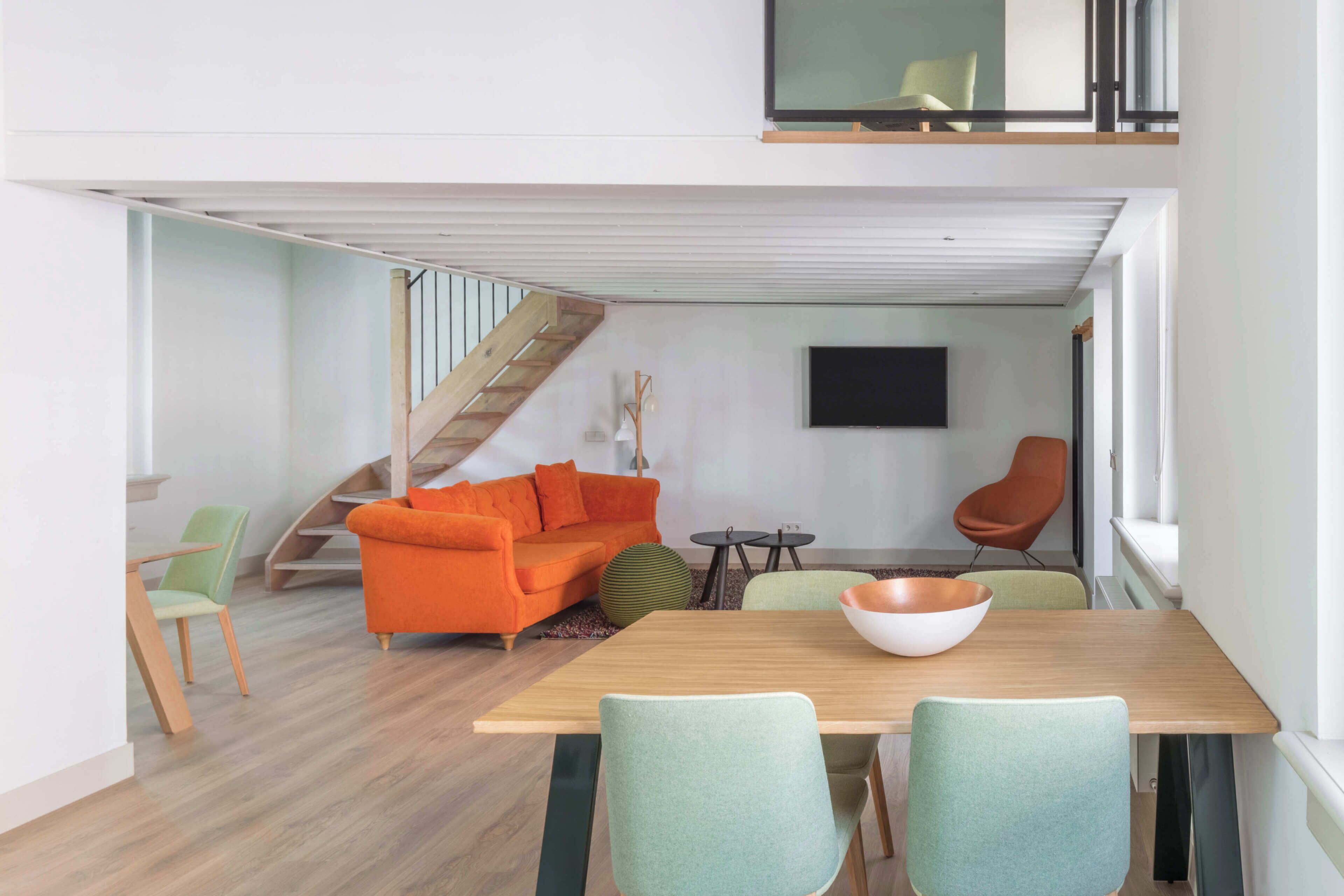 Un salon moderne avec une table à manger en bois, deux chaises vertes, un canapé orange, un fauteuil marron, un escalier et un téléviseur à écran plat fixé au mur.