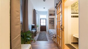 Un estrecho pasillo que da a un dormitorio con una cama, un escritorio con una computadora, una planta en maceta y un armario de madera.