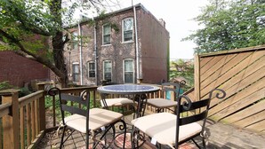 Une terrasse extérieure avec une table ronde et quatre chaises, entourée d’une plateforme en bois et, au fond, d’un bâtiment en briques.