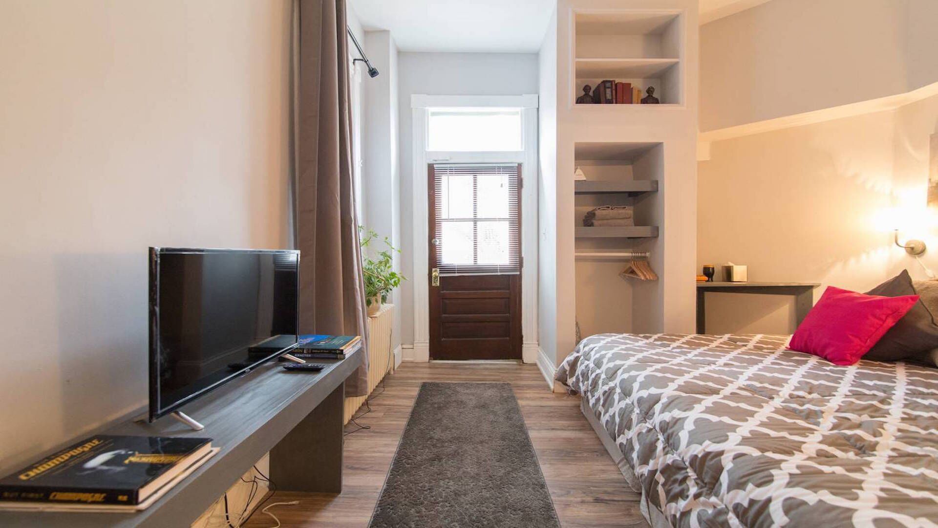 Quarto de hotel pequeno com cama, televisão, escrivaninha com livros, janela com persianas e uma pequena planta.