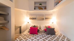 Un dormitorio compacto con una colcha estampada, dos lámparas de pared, una obra de arte con vista a la ciudad, una estantería con libros y una planta, y un banco de madera con toallas.