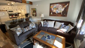 A modern living room with a gray sofa, a coffee table, and a dining area with a kitchen in the background. - Walk to StoryLand! Updated Condo! WiFi/Golf/AC/Pool - New Pool 2026 (Glen)