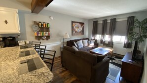 Una sala de estar acogedora con un sofá marrón, una mesa de centro y un comedor con mantel estampado. Se aprecia una estantería con objetos coloridos y un mapa en la pared.