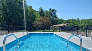 A rectangular swimming pool with metal handrails, surrounded by a black fence, trees, and a building in the background. - Walk to StoryLand! Updated Condo! WiFi/Golf/AC/Pool - New Pool 2026 (Glen)