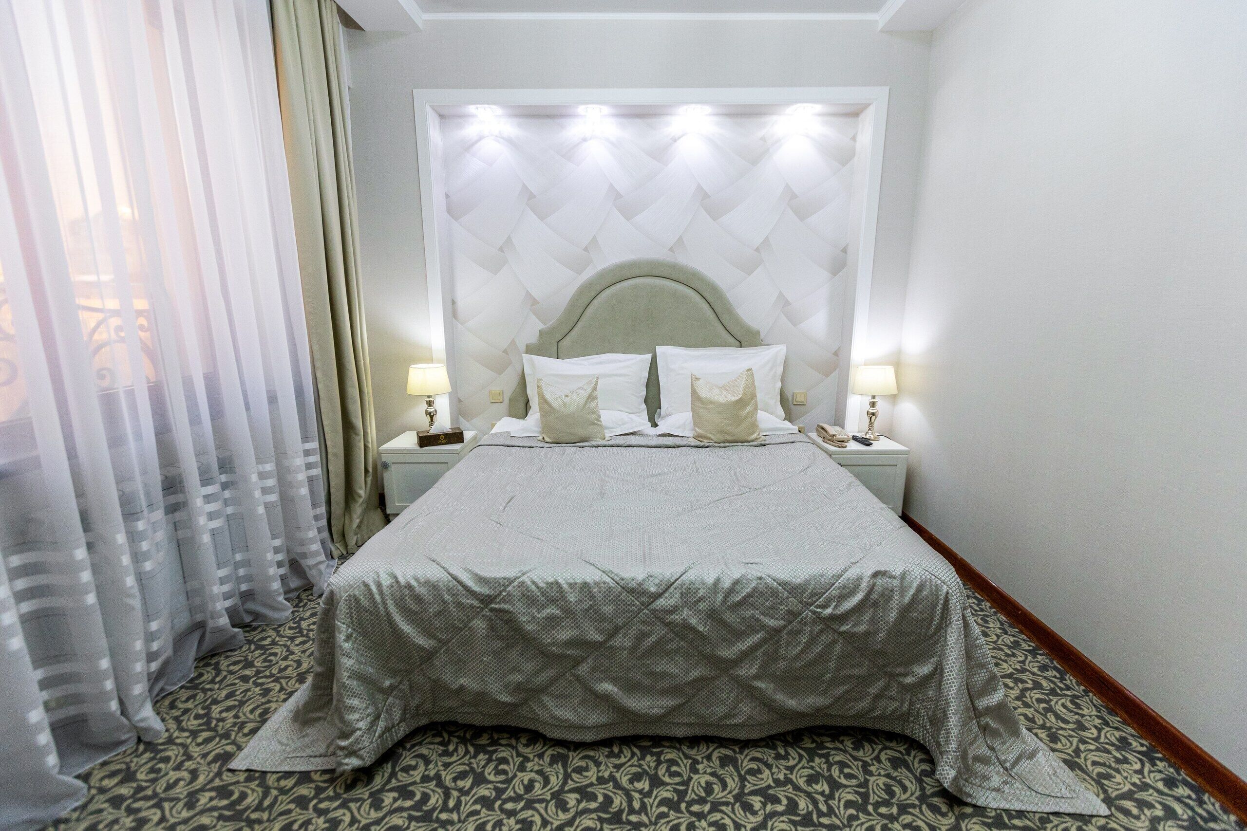 A hotel room with a large bed, a headboard with a geometric pattern, two bedside tables with lamps, and a patterned carpet.