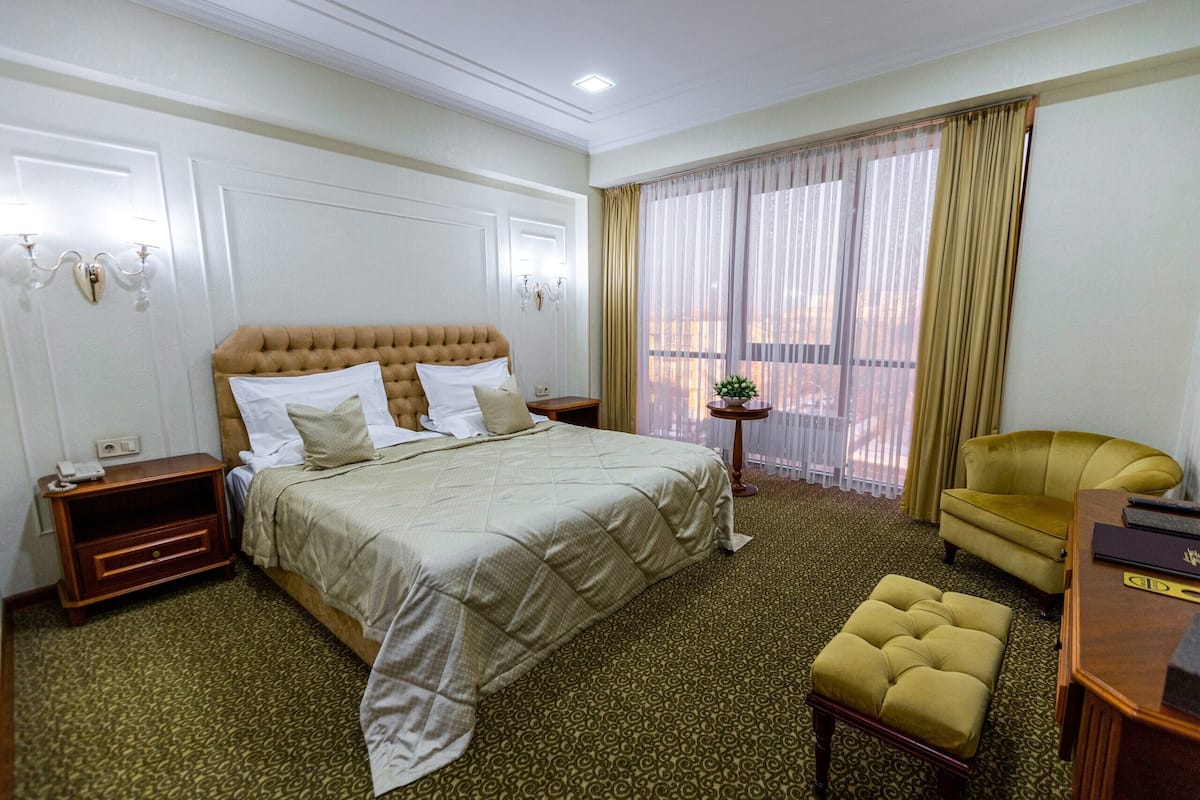 A hotel room with a large bed, a small round table, a yellow armchair, and a wooden desk.