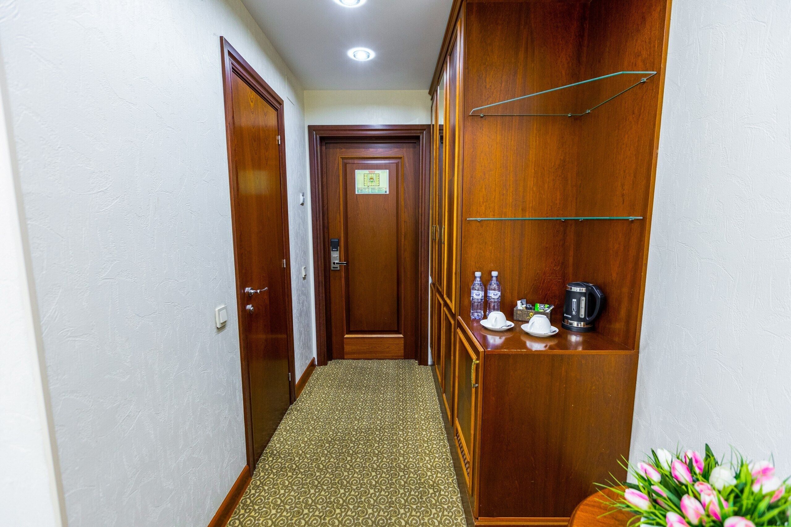 A hallway with a patterned carpet, wooden doors, a shelf with water bottles and a coffee maker, and a small plant.