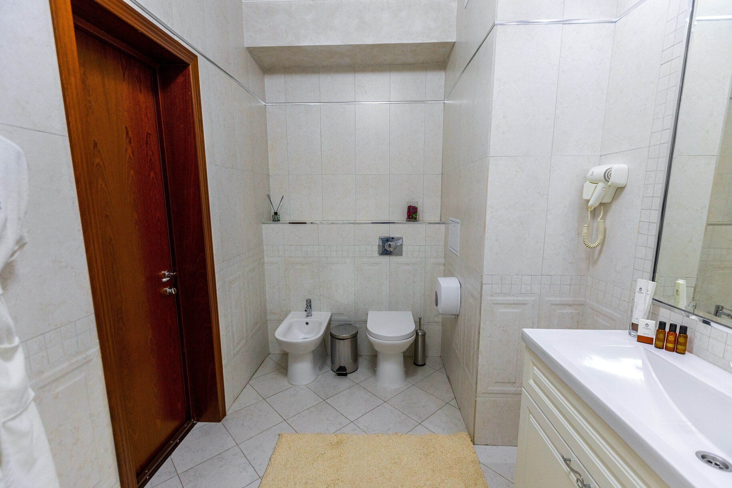 Un baño con puerta de madera, un bidé, un inodoro, un cubo de basura, un perchero para toallas y un lavamanos con artículos de aseo.