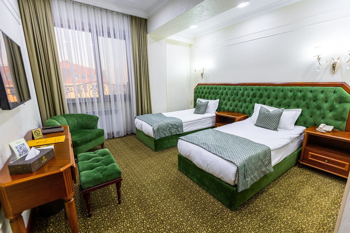 A hotel room with two beds, a green armchair, a wooden nightstand, and a small table with a tissue box and a phone.