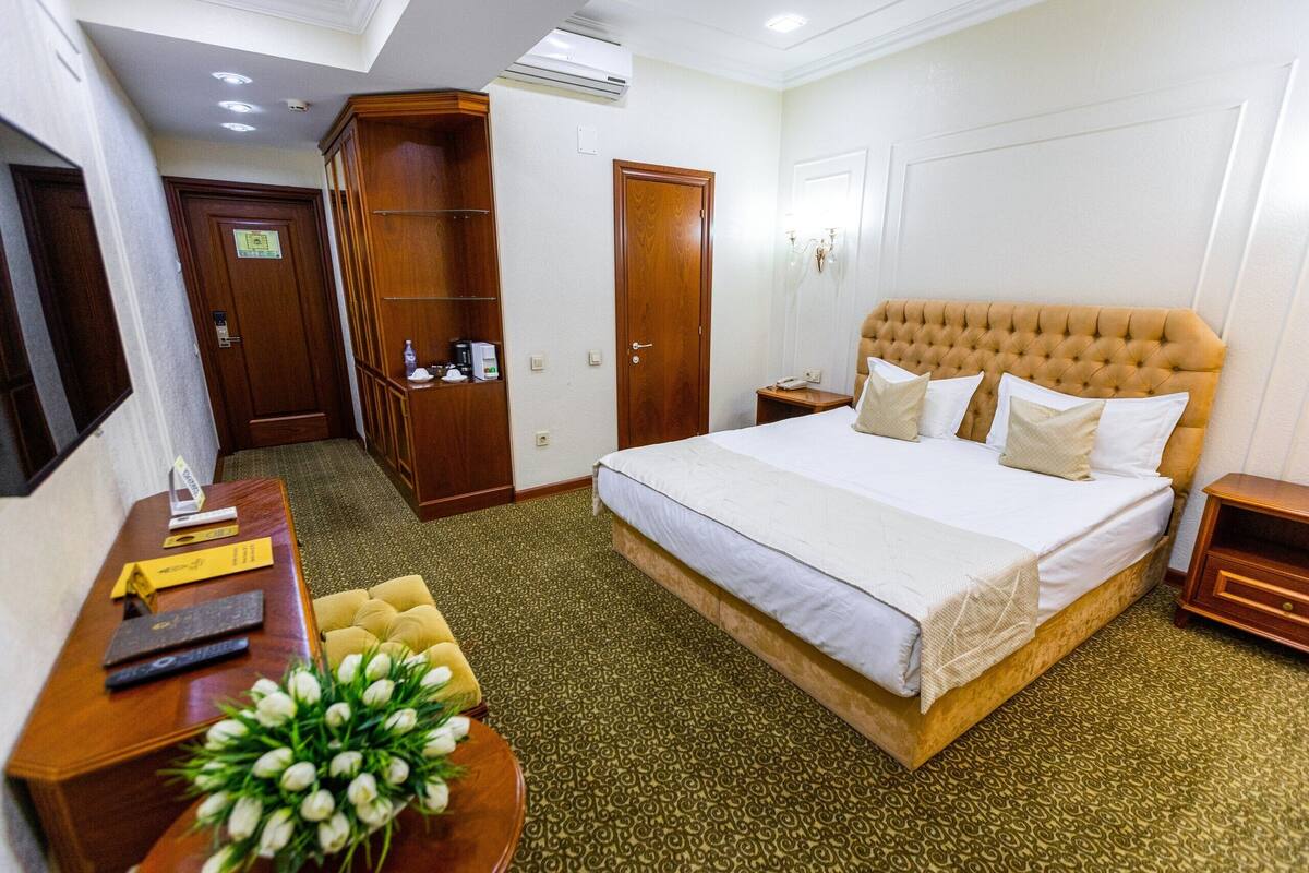 A hotel room with a large bed, a wooden wardrobe, a desk with a telephone, and a small bedside table.