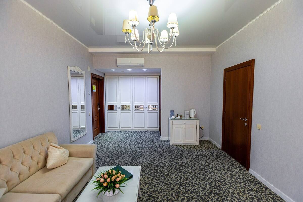A hotel room with a beige sofa, a small table with a vase of flowers, a chandelier, a wardrobe, a cabinet, and a door.